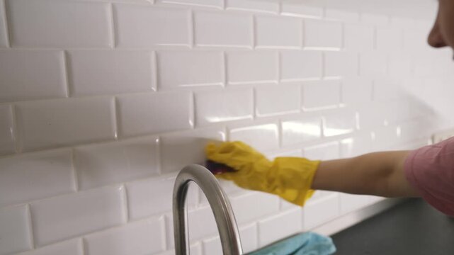Woman wearing yellow gloves cleans white subway tiles on kitchen wall with sponge near faucet