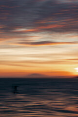 Obraz premium Orange and pink hues fill the sky over a blurred figure on a serene ocean at sunset. Mui Ne, Vietnam