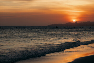 Obraz premium Sun sets over a tranquil ocean with distant hills silhouetted against an orange sky. Mui Ne, Vietnam