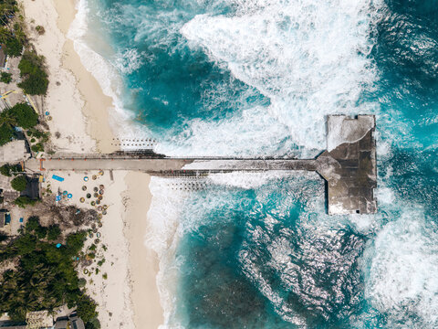 Aerial view of a concrete pier extending into vibrant blue ocean waves at a sandy beach. Ly Son island, Vietnam