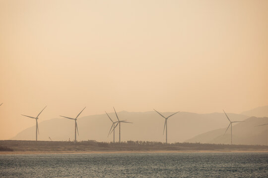 Wind turbines on a hazy coastal landscape with mountains in the background at dusk.