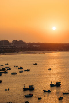 A serene sunset over a bay with numerous boats and distant modern buildings under an orange sky.