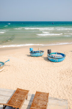 Wooden lounge chairs overlook a sandy beach with three round blue boats by the turquoise sea.
