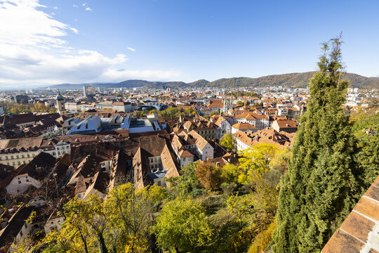 View of the historic old town with red-tiled rooftops, the modern Kunsthaus Graz building, and rolling hills under a blue sky Graz, Styria, Austria.