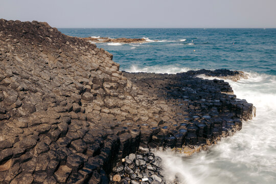 Basalt columns on a rugged coastline with waves crashing against them under a clear sky.