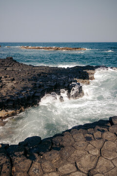 Rugged basalt columns meet crashing ocean waves under a clear blue sky.