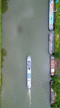 Aerial view of a long white cargo barge with solar panels navigating a canal with moored boats and green banks in Anderlecht, Brussels, Belgium.