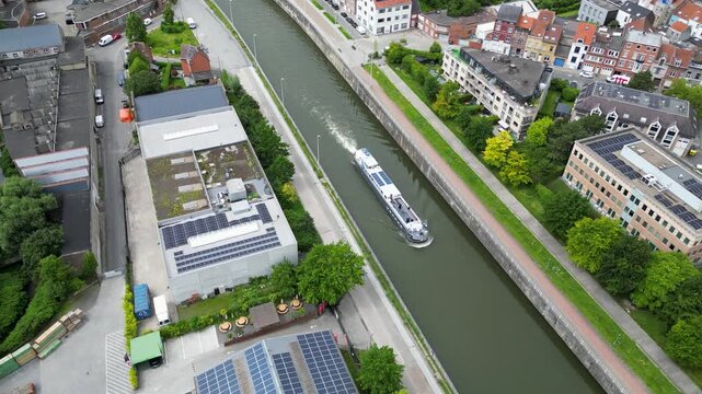 Aerial view of a cargo barge navigating the canal past industrial buildings with solar panels and residential areas in Anderlecht, Brussels, Belgium.