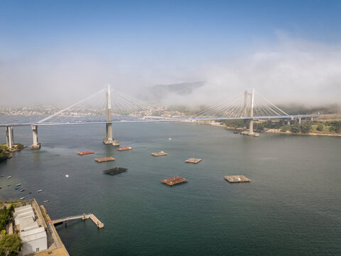 Aerial view of a suspension bridge spanning a wide river with misty hills in the background.