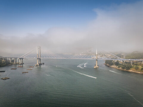 A suspension bridge stretches across a wide river under a partly cloudy blue sky.