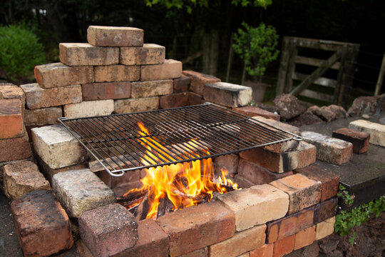 An outdoor brick barbecue with a metal grill is burning wood flames in a garden setting.