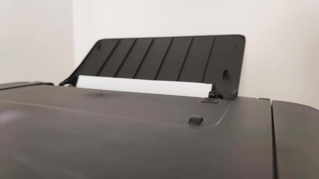 Close up of automated printing process in office. White blank A4 paper sheet is feeding into black multifunction machine to start automated printing process of corporate business documents.