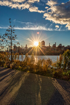 Sun setting behind a city skyline casting long shadows beside a scenic river path. New York, USA
