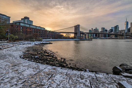 Snow-dusted pathway curves by a river with a city skyline and a bridge at sunset. New York, USA