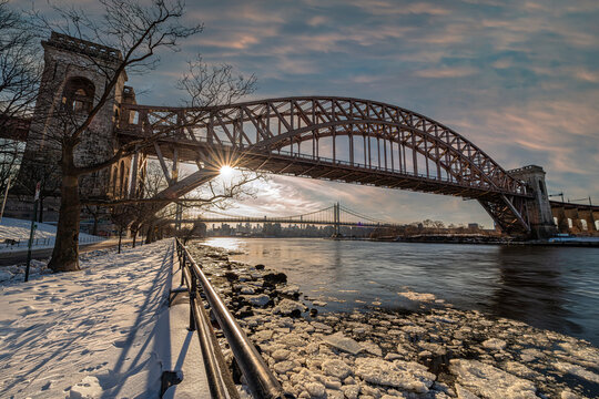 Snowy river bank with a large, arched bridge under a dramatic sunset sky. New York, USA
