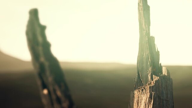 Wooden posts rise from a field, set against a soft sunset backdrop. The sky glows while distant hills create a silhouette effect. The scene feels natural and simple.