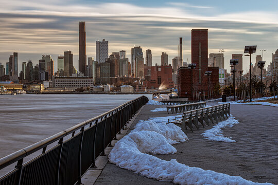Snow-lined walkway along river with city skyline under cloudy sky during daylight. New York, USA