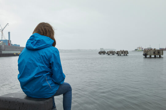 Person in blue jacket sitting by a waterfront on a cloudy day with cranes and buildings in view.  
