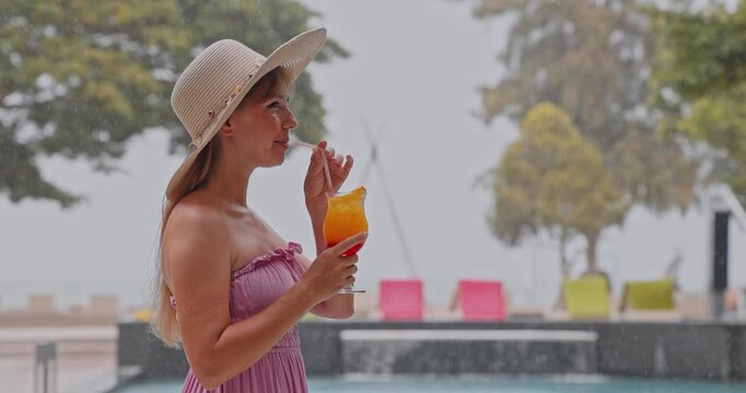 Woman wearing straw hat sipping orange cocktail, rainy tropical resort poolside with lounge chairs, leisure travel, summer holiday weather