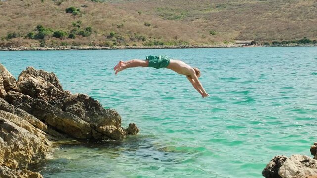 Slow motion shot of man diving from coastal rocks into turquoise sea. Summer action scene shows side view jump above clear water, rocky shore and sunlit hills in bright summer daylight.