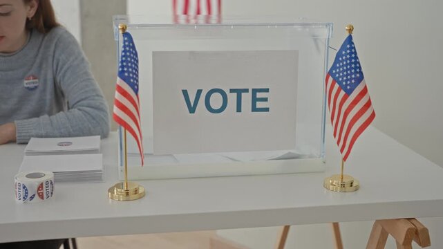 Woman writing on clipboard with pen at voting table beside clear ballot box and small us flags in a polling building; patriotic civic duty.