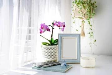 Vertical blank picture frame mockup with a pink orchid, hanging plant, and notebook on a bright windowsill. Nature-inspired cozy home office workspace, airy curtains, soft morning light. © Laura Pashkevich