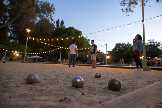 Metal balls rest on sandy court for bocce game at park. People play outdoor game under string lights at dusk. Evening leisure activity with friends.