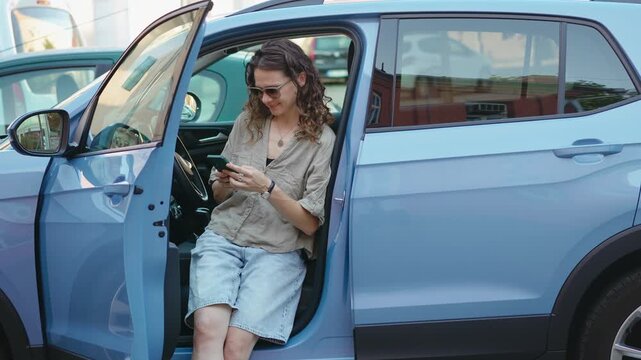 Young female driver using a smartphone while standing next to a parked car on a city street in Europe, representing modern mobility and connected lifestyle.