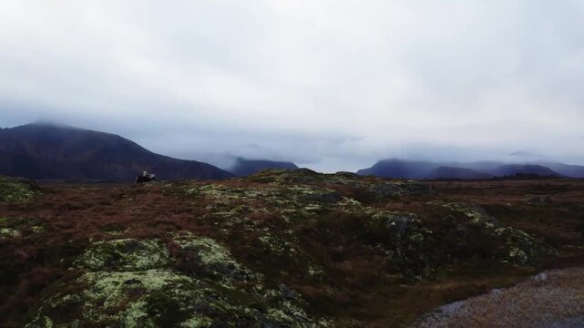 Extreme wide shot of a woman riding a dark horse across rocky valley terrain, stopping to gaze at mist-covered mountains under a cloudy sky, creating a dramatic, cinematic wilderness scene.