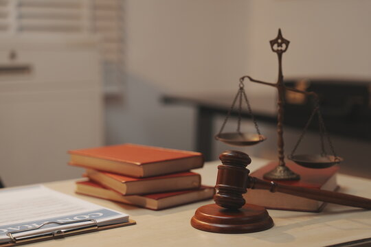 A dramatic, selective-focus close-up shot of a wooden judge's gavel being struck down onto its sound block. In the blurred background, a lawyer in a dark suit sits at a polished desk.  