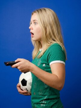 Stunned blonde spectator channel surfing with remote while holding football on blue background in sharp studio focus for cable tv marketing