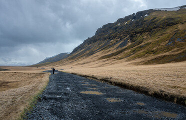 Hiker trekking along a desolate gravel f road in a vast valley, surrounded by mountains with patches of snow and dry grass. Icelandic landscape, Iceland © Michalis Palis