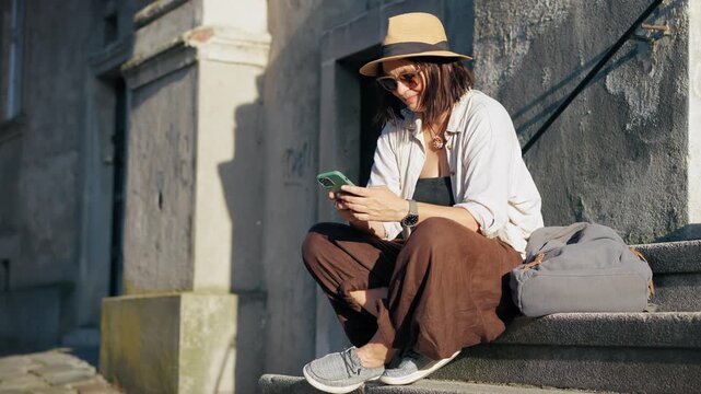 Female traveler in a hat looking at her smartphone while sitting on the steps of a historic building in a European city during a beautiful sunset.