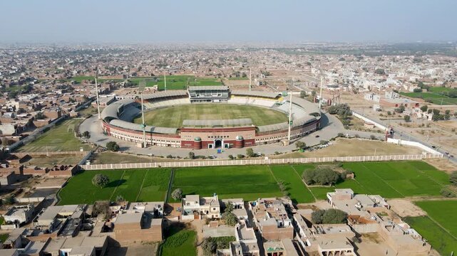 Aerial view of Multan Cricket Ground with its circular stands and green pitch overlooking the city and agricultural fields in Multan, Punjab, Pakistan.