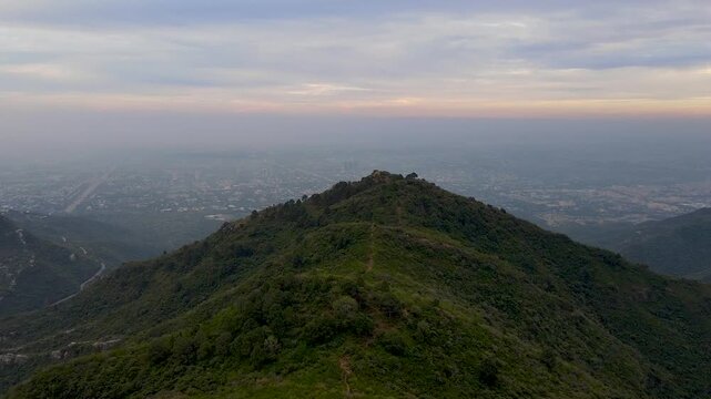 Aerial view of the lush green Margalla Hills ridge overlooking the hazy city skyline during twilight in Islamabad, Islamabad Capital Ter, Pakistan.