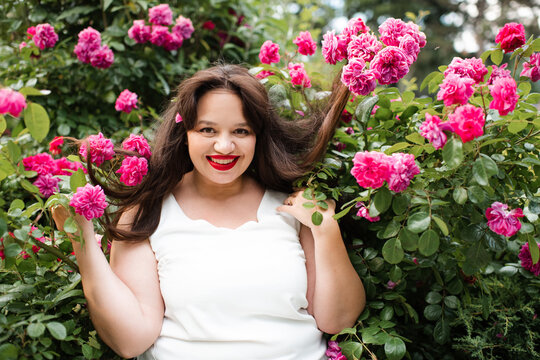 Happy young adult woman 30-35 year old with red lipstick make up and long hair in blooming rose bushes with flowers in garden outdoors close up. Summer vacation season. Healthy lifestyle.