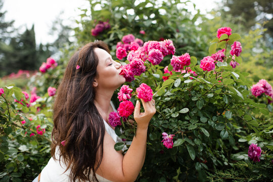 Beautiful young adult woman 30-35 year old with long hair in smelling rose flowers in garden outdoors close up. Summer vacation season. Healthy lifestyle.