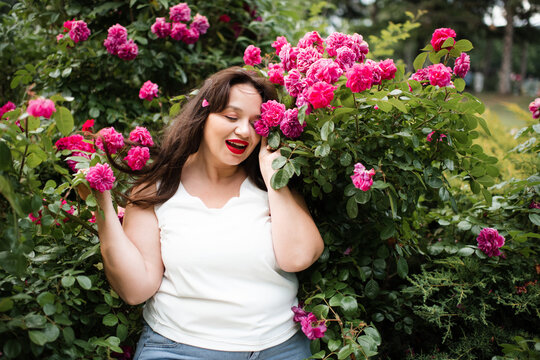 Smiling young adult woman 30-35 year old with red lipstick make up and long hair posing in blooming rose flowers in garden outdoors close up. Summer vacation season. Healthy lifestyle.