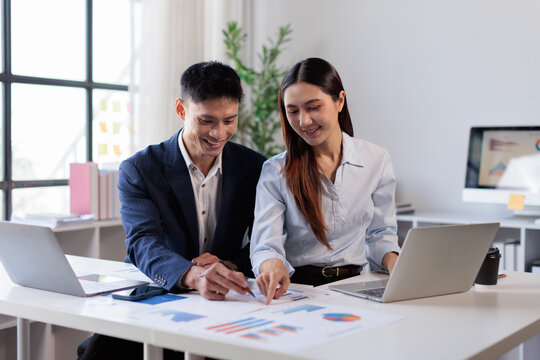 Two asian confident business people point to graphs and charts to analyze market data, balance sheet, account, net profit to plan new sales strategies to increase production capacity Financial and Acc