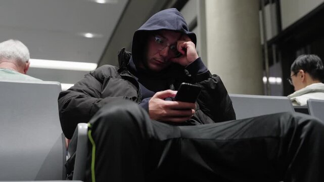 Tired man wearing hoodie and jacket scrolling smartphone while waiting in airport departure lounge with other passengers