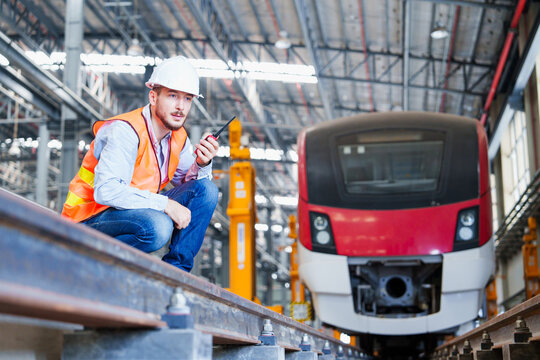 Industrial railway technician performs detailed inspection of electric train within expansive workshop utilizing handheld radio and standard safety apparel &ndash; white helmet, orange vest, light-colored s