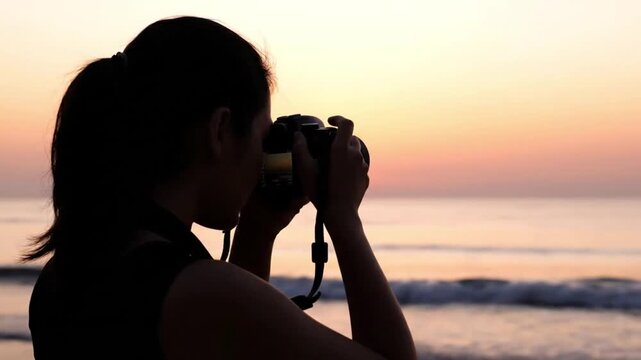 Silhouette of female travel photographer using digital dslr camera on tropical beach during vibrant orange golden hour sunrise