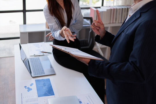 Two asian confident business people point to graphs and charts to analyze market data, balance sheet, account, net profit to plan new sales strategies to increase production capacity Financial and Acc
