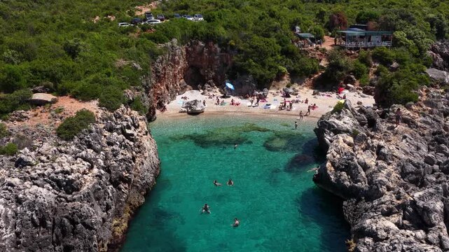 Aerial view of Aquarium Bay featuring turquoise waters, rocky cliffs, and people relaxing on the small sandy beach surrounded by lush greenery Himara, Qarku i Vlores, Albania.