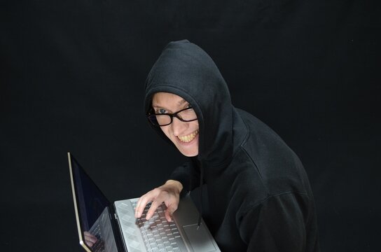 Sneaky cyber criminal in glasses and black hoodie looking at camera with evil smile while hacking laptop.