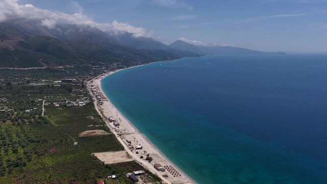Aerial view of the turquoise Ionian Sea coastline with a sandy beach and green mountains under a blue sky in Llogara, Vlore County, Albania.