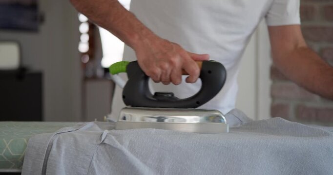 Ironing Shirt at Home: A man's hands carefully guide an iron over a patterned shirt, smoothing out wrinkles on an ironing board, with a brick wall in the background.
