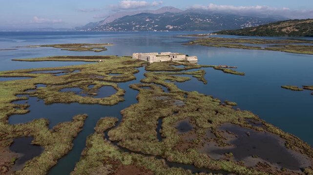 Aerial view of Ali Pasha Castle surrounded by marshy wetlands and calm blue water with mountains in the background in Mahala, Qarku i Vlores, Albania.
