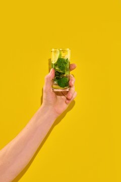 Hand holding mojito cocktail on yellow background. Concept of summer resort drinks, brunch culture, mocktail market, cocktail menu, and social media content.