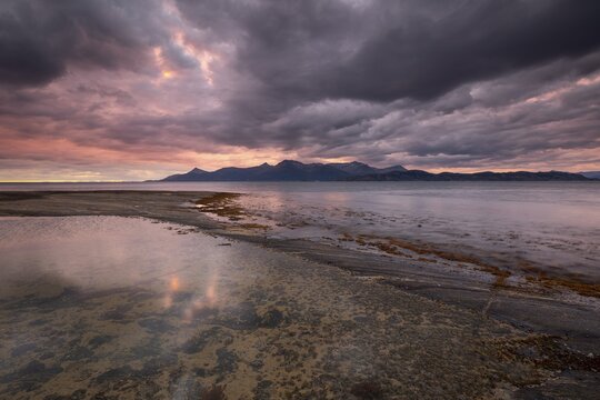The Vestfjord in Nordland, Norway, offers a dramatic atmosphere near Bod&oslash;. Dark clouds are illuminated by the setting sun in shades of red and yellow. The rugged cliffs and mountains are reflected in the quiet fjord, which offers a breathtaking view of Lofoten